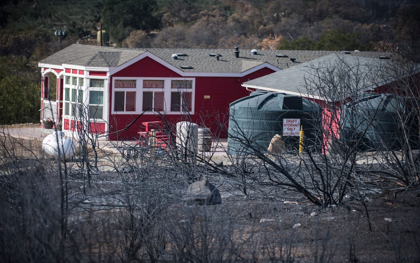A red house still standing in burned brush after the 2017 Thomas Fire in California, its cleared yard, water tank and propane tank untouched. Photo: Stuart Palley for the US Forest Service.
